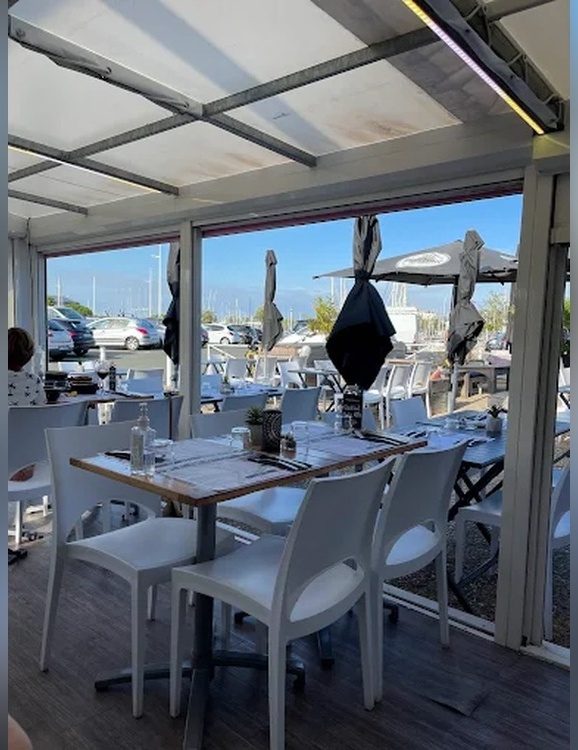 La terrasse couverte et ses tables dressées ; au loin, derrière le parking, les mâts du port des Minimes